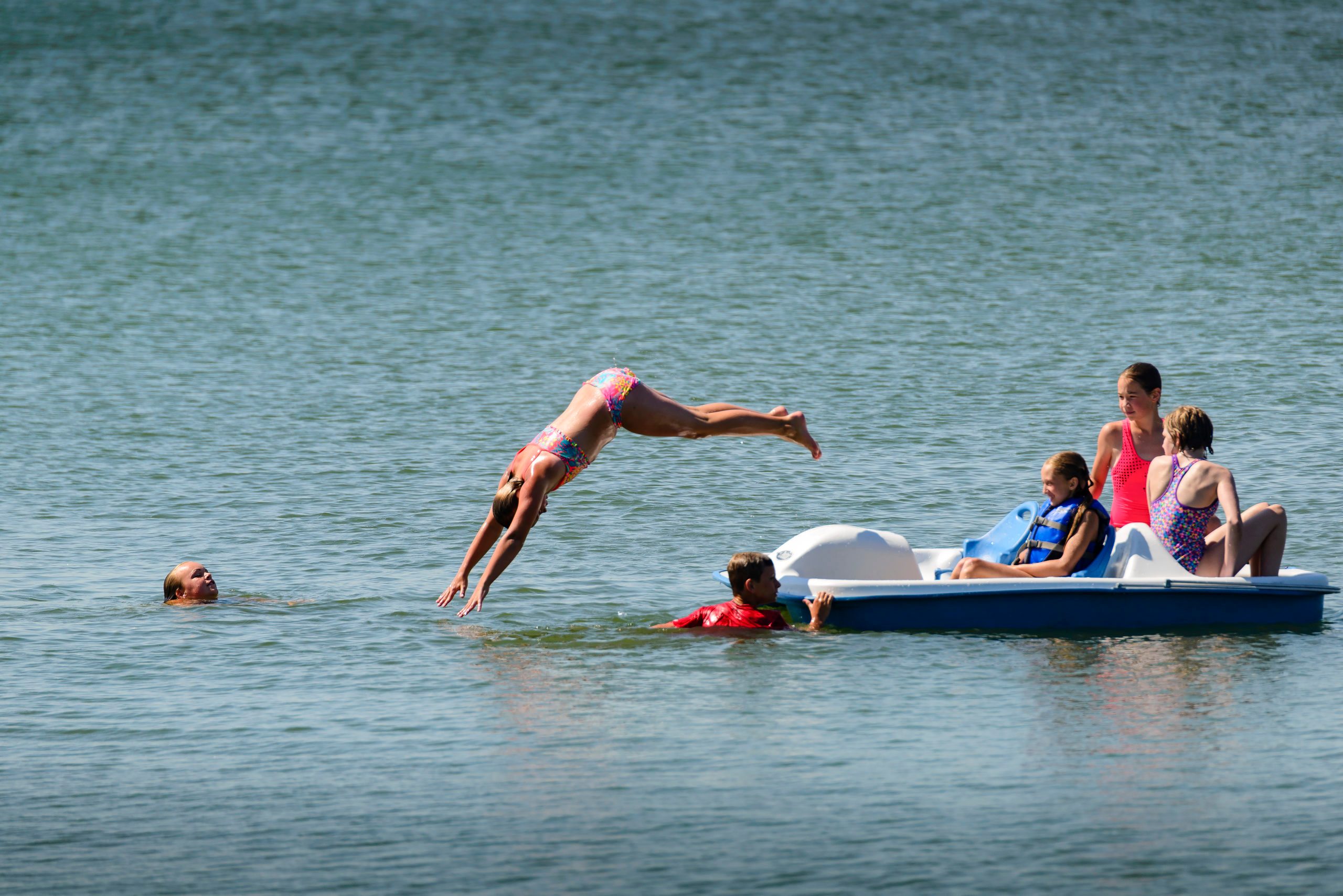 A person dives into the water, near a pedal boat with people, in a large lake on a clear day.