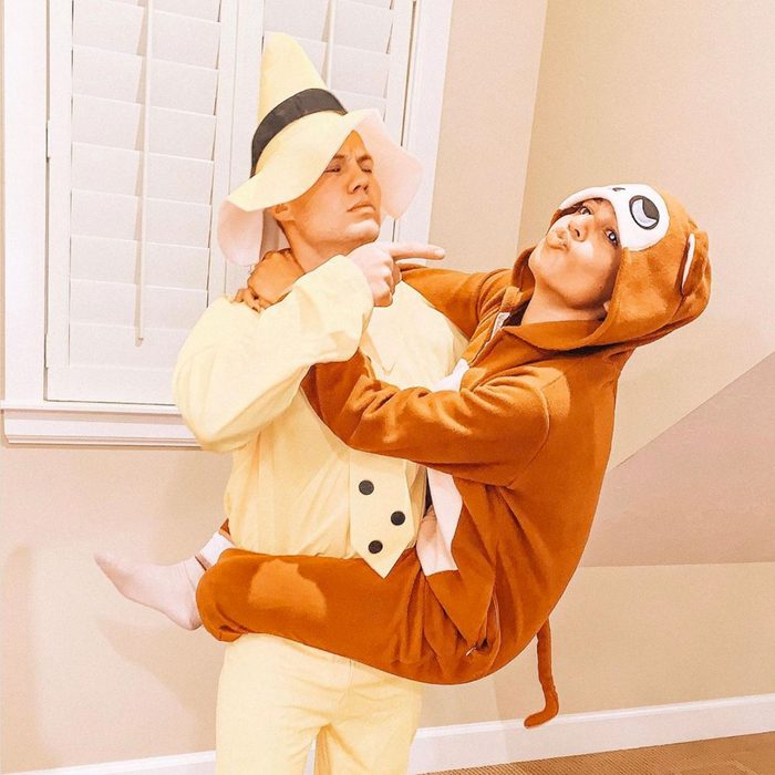 Person in yellow outfit lifts person in brown monkey costume indoors against a window with shutters.