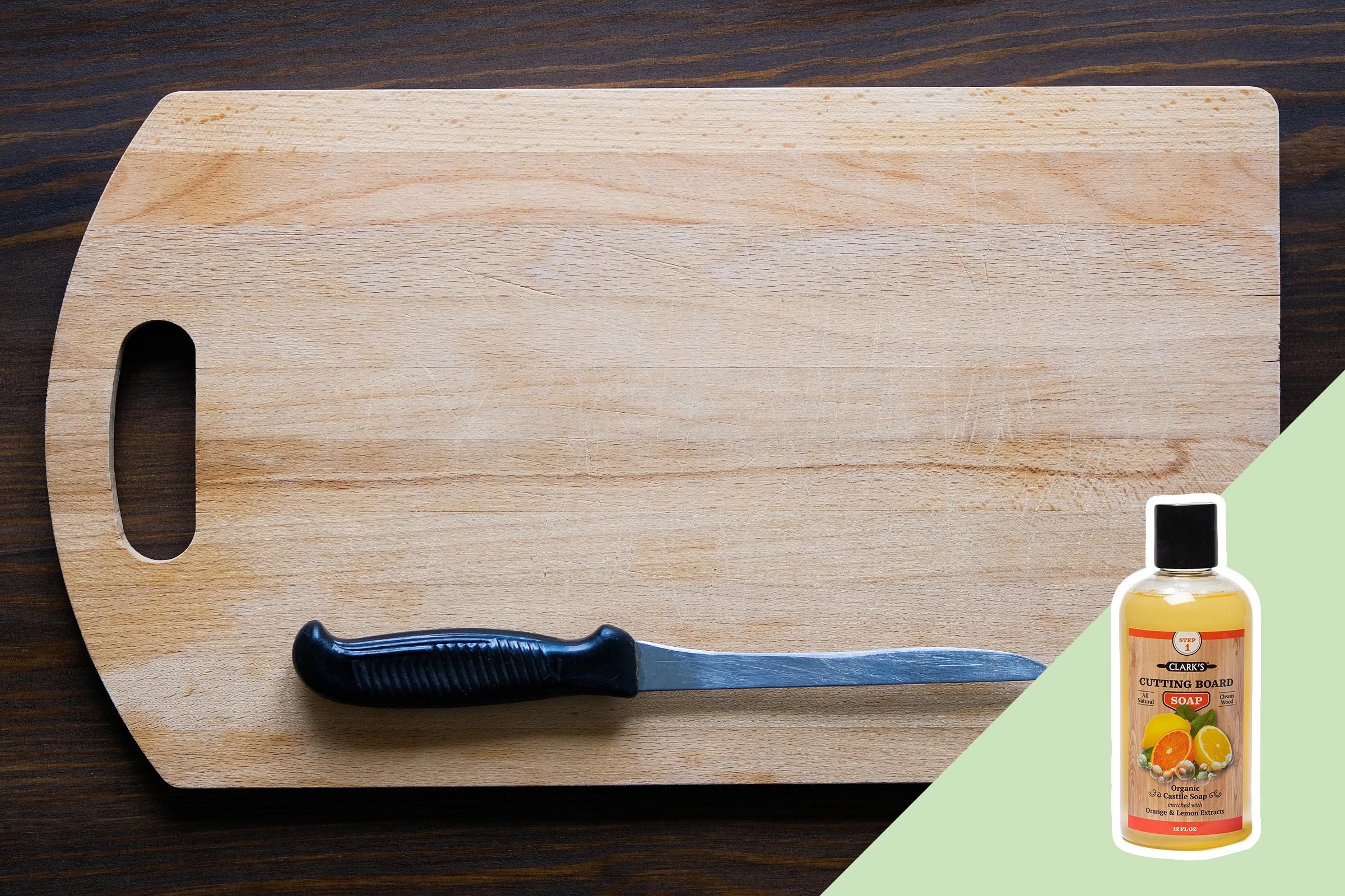 A wooden cutting board with a black-handled knife rests on a dark surface, accompanied by a bottle of cutting board soap.