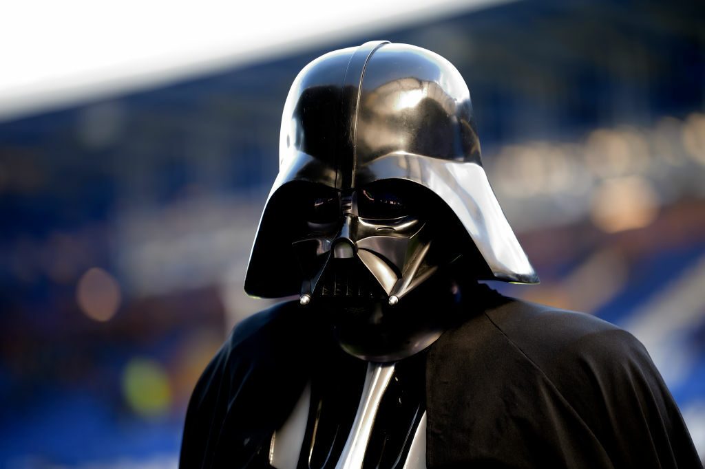 A person wears a shiny black helmet and cape, standing against a blurred, colorful background.