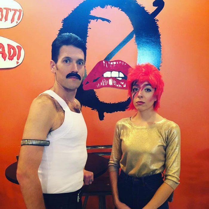 Two people pose in costumes, one resembling a musician with a mustache and white tank top, the other with red hair and gold top, against an orange wall.