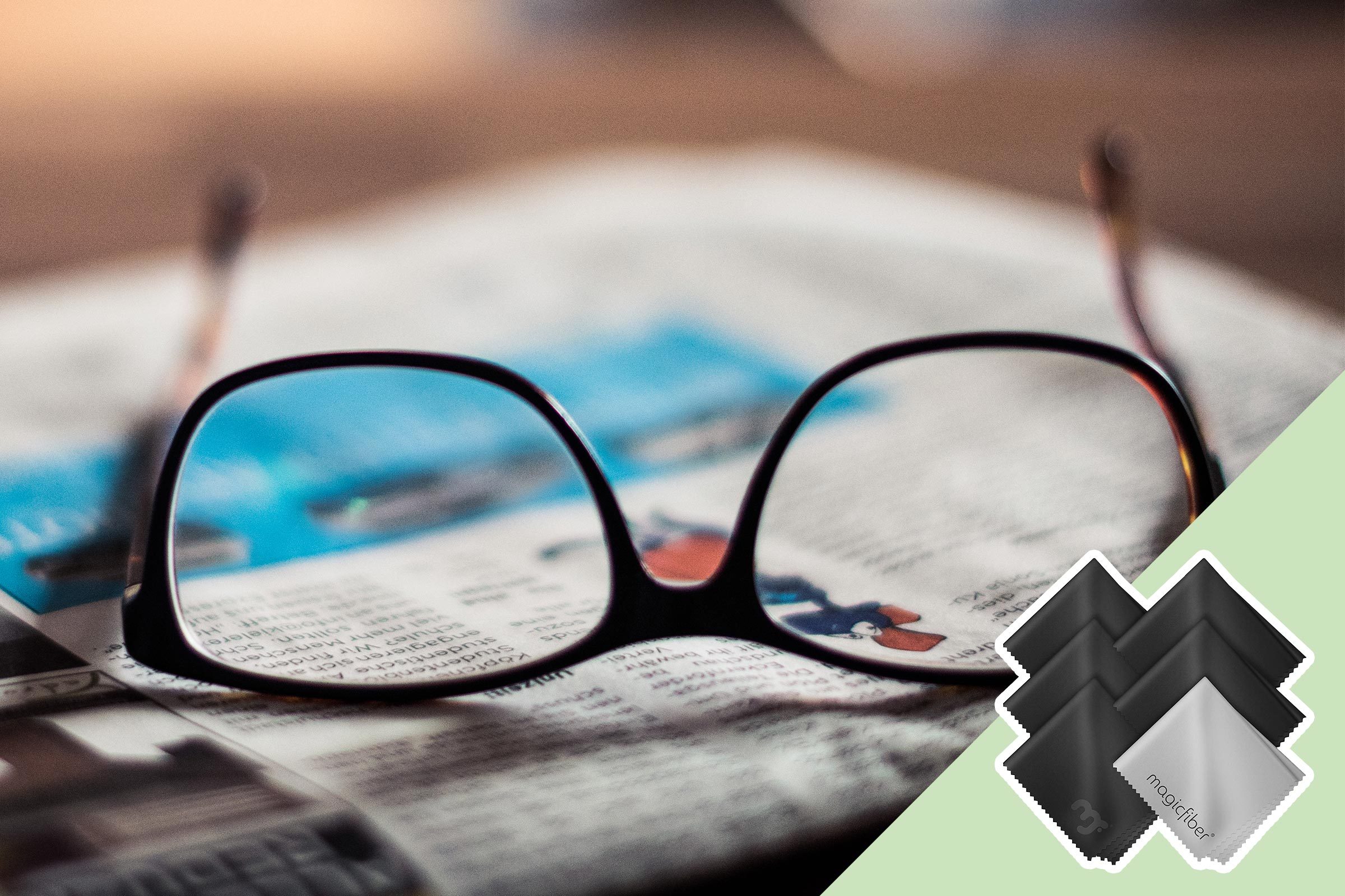 Glasses resting on an open newspaper; nearby, five folded cleaning cloths, one labeled "magicfiber," lie on a green surface.