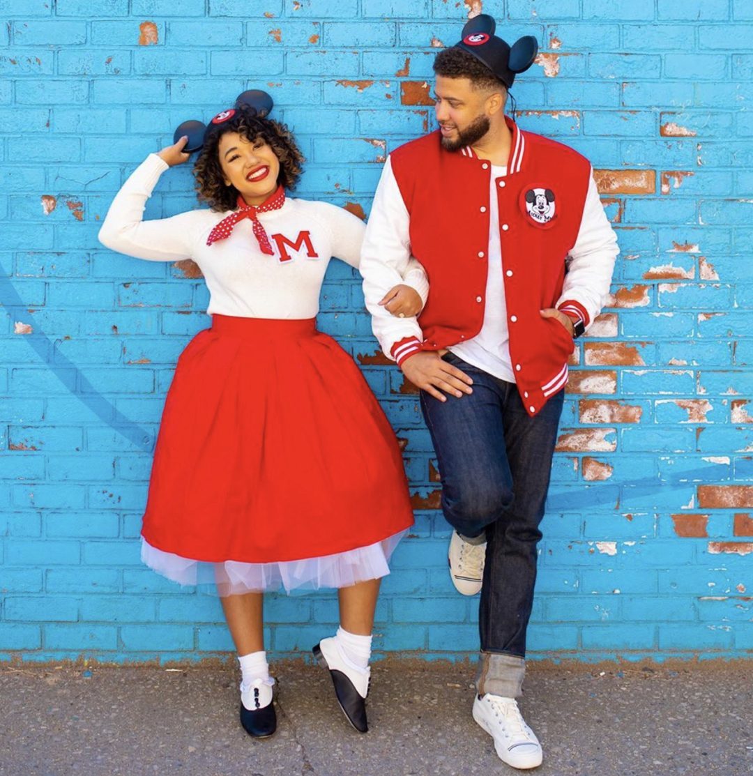 Two people wearing vintage-style outfits, smiling, and posing playfully against a bright blue brick wall.
