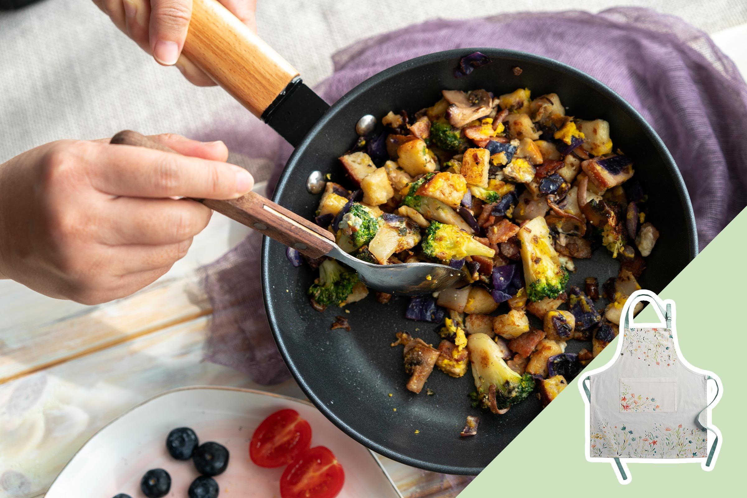 Hands stir-frying vegetables in a pan; a plate with blueberries and tomatoes nearby. An apron is pictured against a light green background.