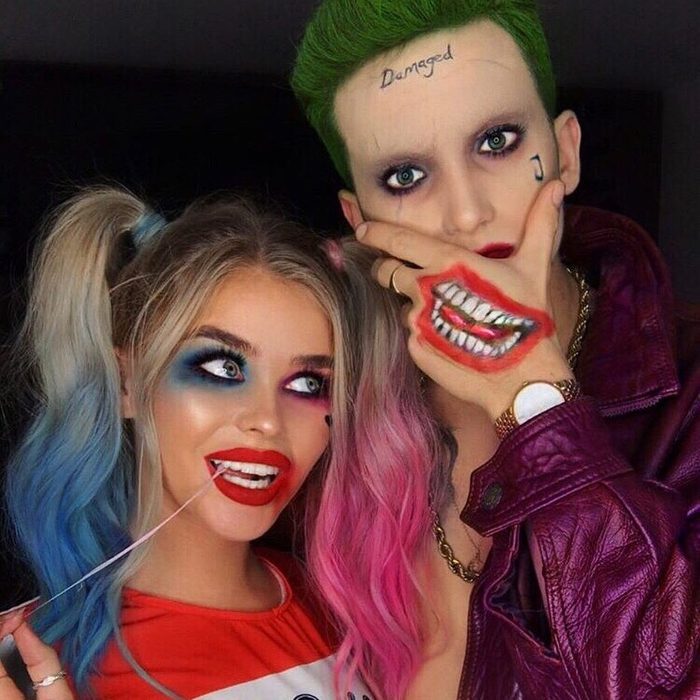 Two people in vibrant makeup pose playfully; one chews gum and the other covers a tattooed smile with their hand, wearing purple clothing.