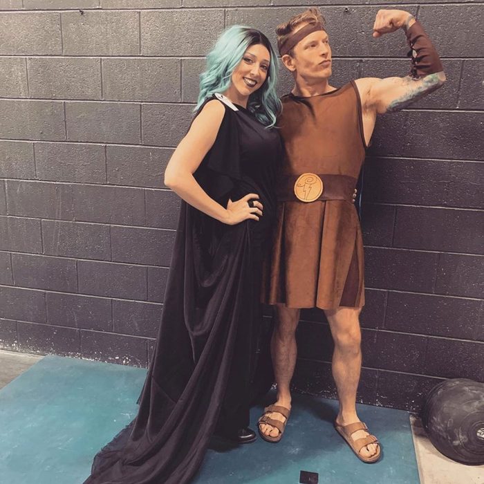 Two people in costumes pose playfully; one flexes in Greek attire, the other in a flowing black dress, against a dark brick wall backdrop.