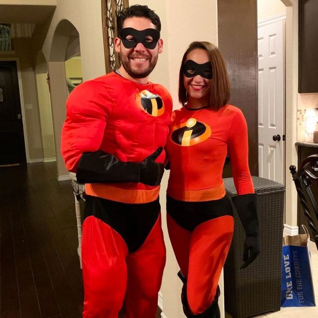 Two people wear superhero costumes, smiling indoors by a hallway. Text on bag reads: 