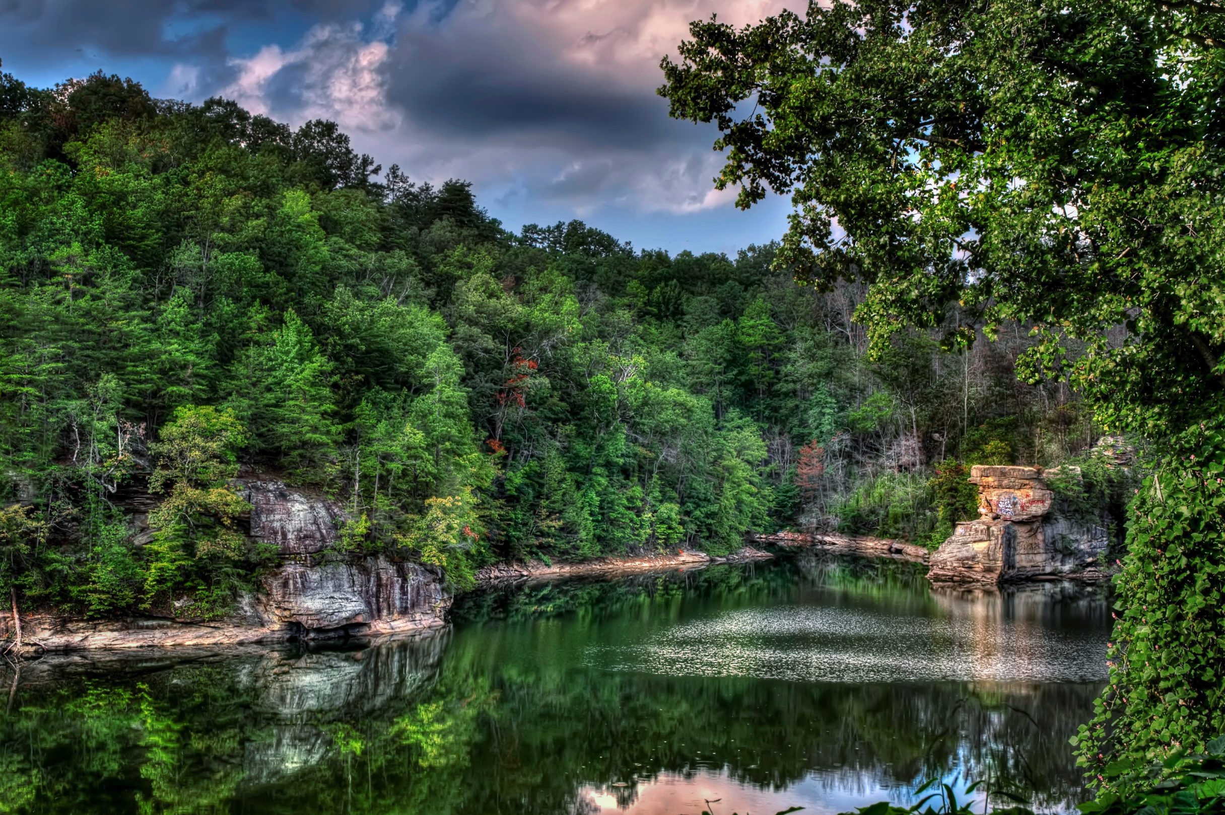 Lake reflects lush green forest and rocky cliffs under cloudy sky, creating a serene and picturesque natural landscape.