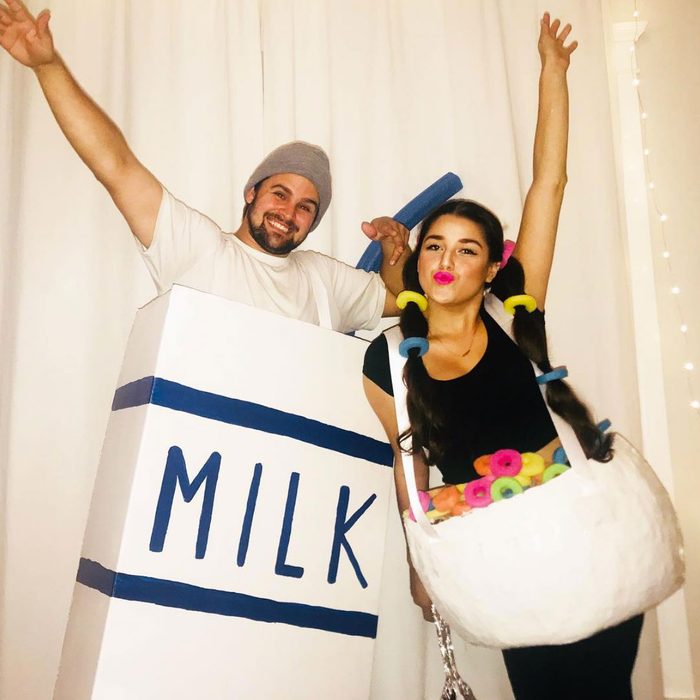 Person dresses as milk carton, smiling, raises arm; another as cereal bowl with colorful loops, poses playfully. White curtain backdrop suggests indoor setting.