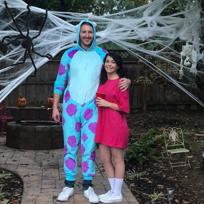 Two people wearing costumes stand smiling; one is in a blue onesie with purple spots, the other in a pink dress, surrounded by Halloween decorations.