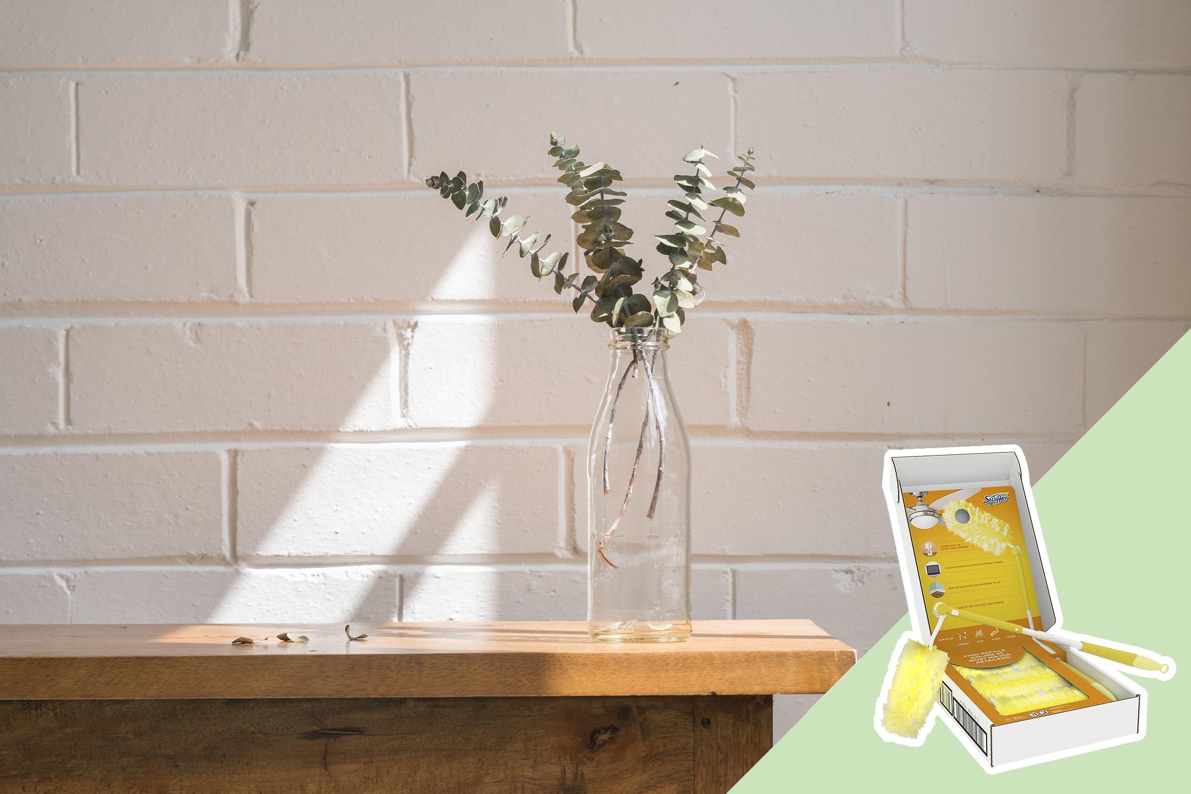 A glass vase holds eucalyptus branches, casting shadows on a wooden surface against a white brick wall; nearby is a cleaning duster in an open box.