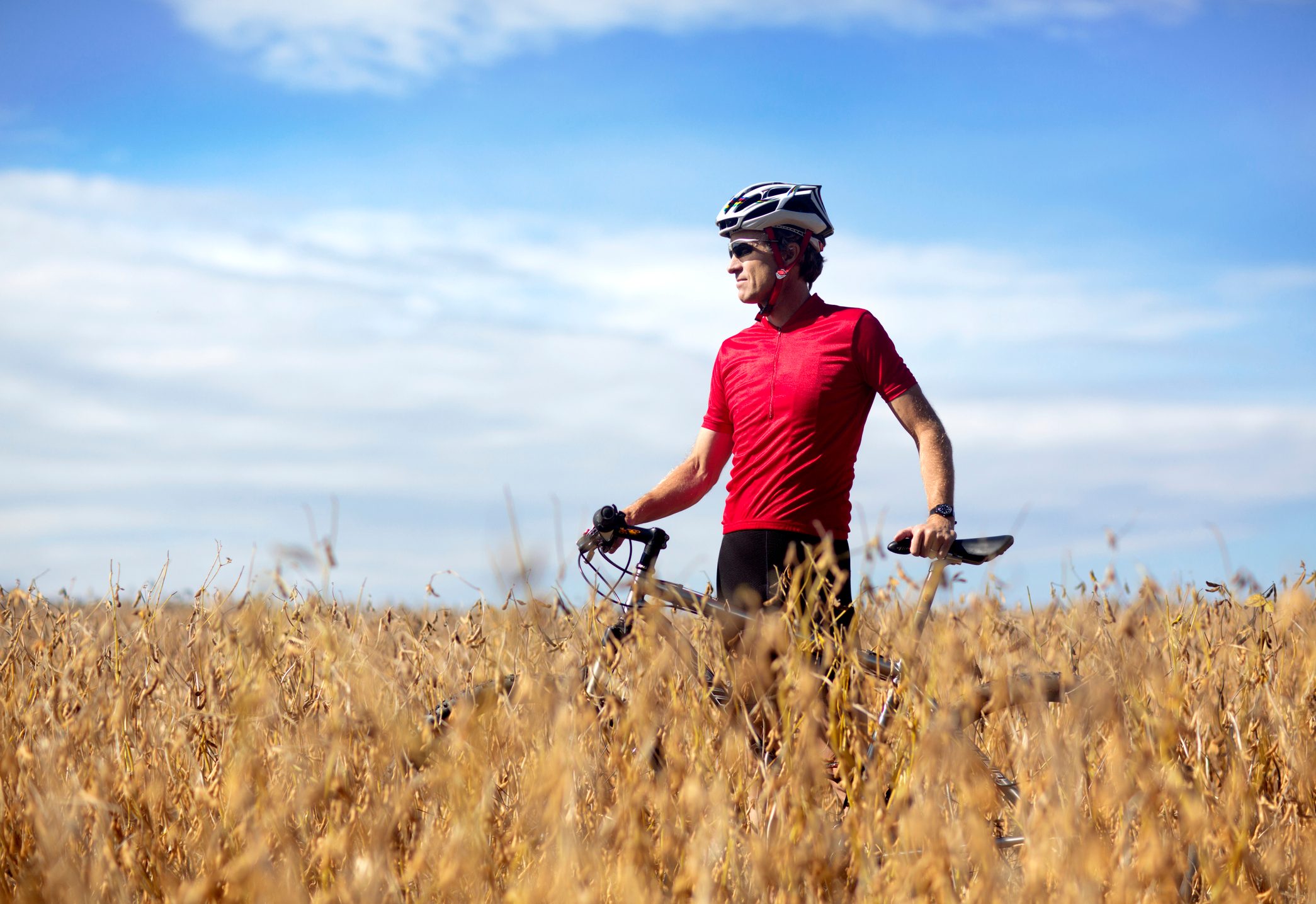 Cyclist stands with a bike, wearing a red jersey and helmet, surrounded by a golden wheat field under a clear blue sky.