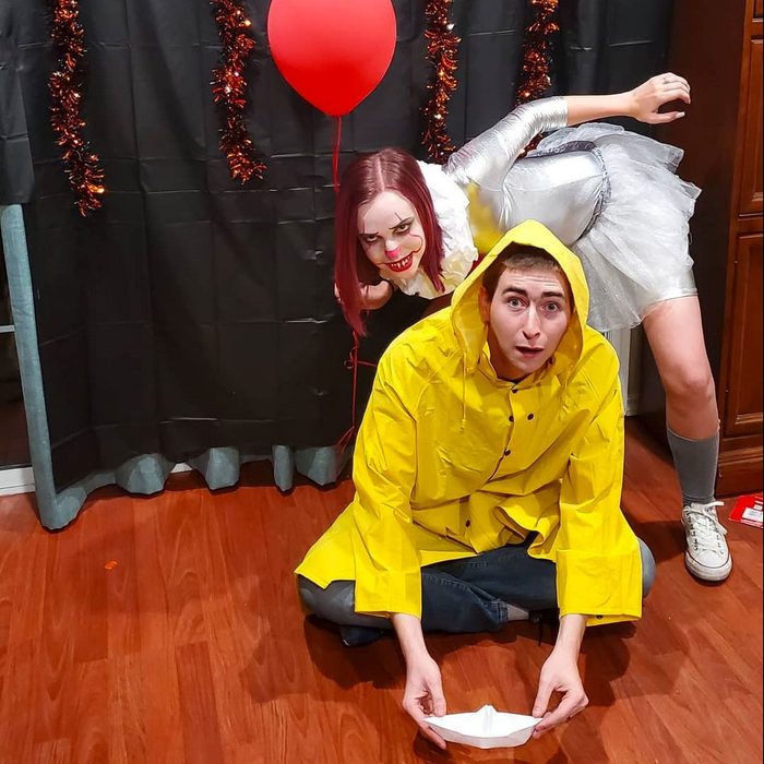 Person crouches in a yellow raincoat, holding a paper boat; clown-like figure behind holds a red balloon, inside a decorated room.