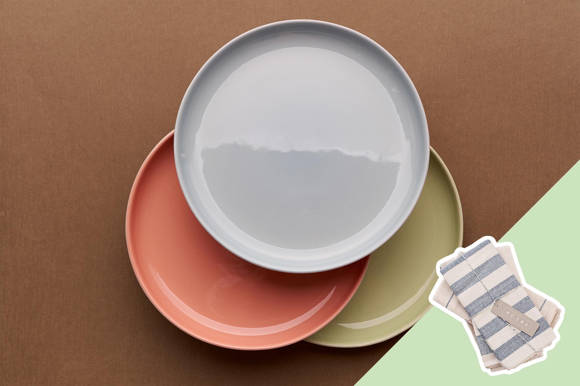 Three overlapping ceramic plates in pastel colors sit on a brown surface; two striped cloth napkins are placed on a green background.