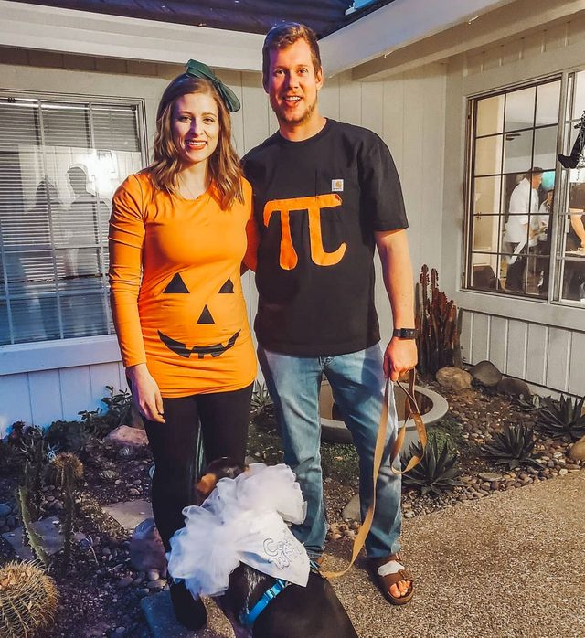 Two people wear Halloween costumes, one as a pumpkin and the other with the pi symbol, beside a dressed dog in a yard.