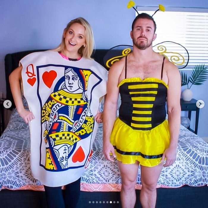 A woman wears a card costume; a man wears a bee outfit. They stand smiling in a bedroom with a bed and plants.
