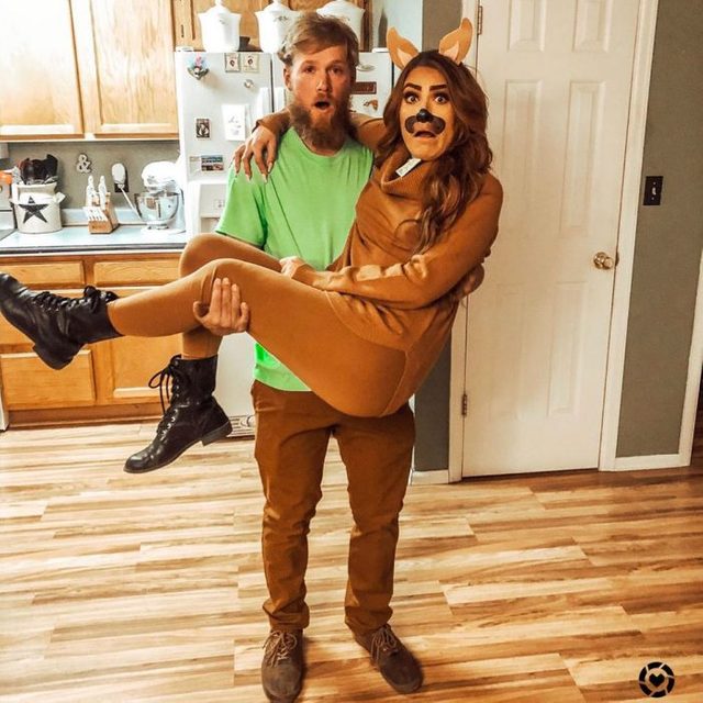 A person in green holds another dressed as a dog, standing on a wooden floor in a kitchen.