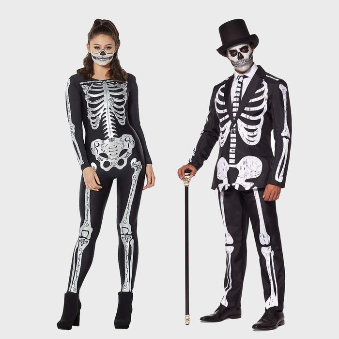 Two people wear skeleton costumes standing together; one with a cane and top hat, the other with a skull mask, against a plain background.