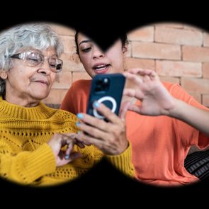Two people use a smartphone together, with one explaining to the other on a couch, framed through binocular-like view.