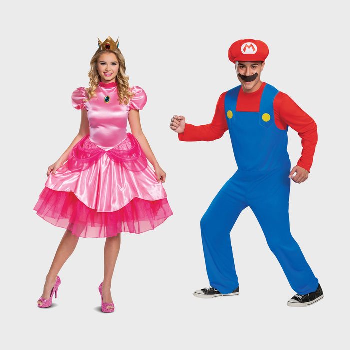 Two people pose in colorful costumes: one in a pink dress and crown, the other in blue overalls and a red hat, against a plain background.