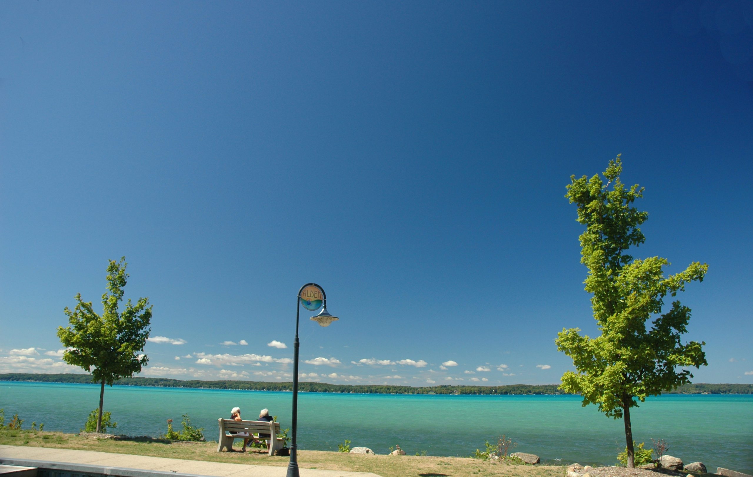 Two people sit on a bench, overlooking a serene blue lake surrounded by grassy banks and trees under a clear sky.