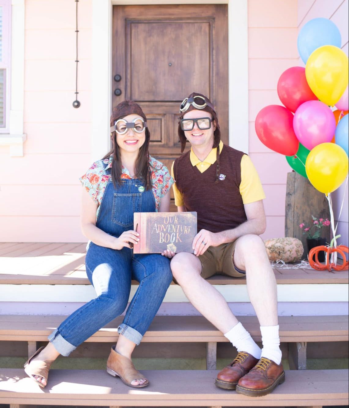 Two people in vintage aviator outfits hold "Our Adventure Book" on porch steps, surrounded by colorful balloons.
