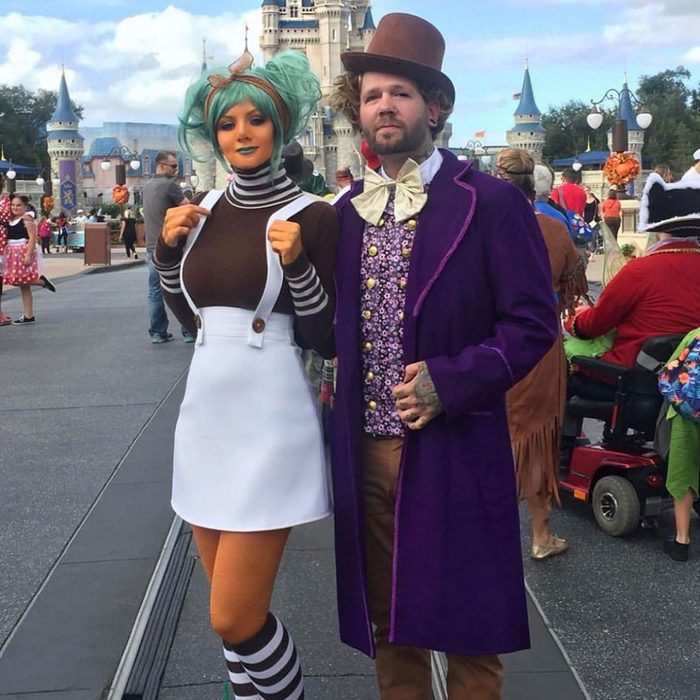 Two people in costumes pose in front of a castle at a theme park, surrounded by crowds and festive decorations.