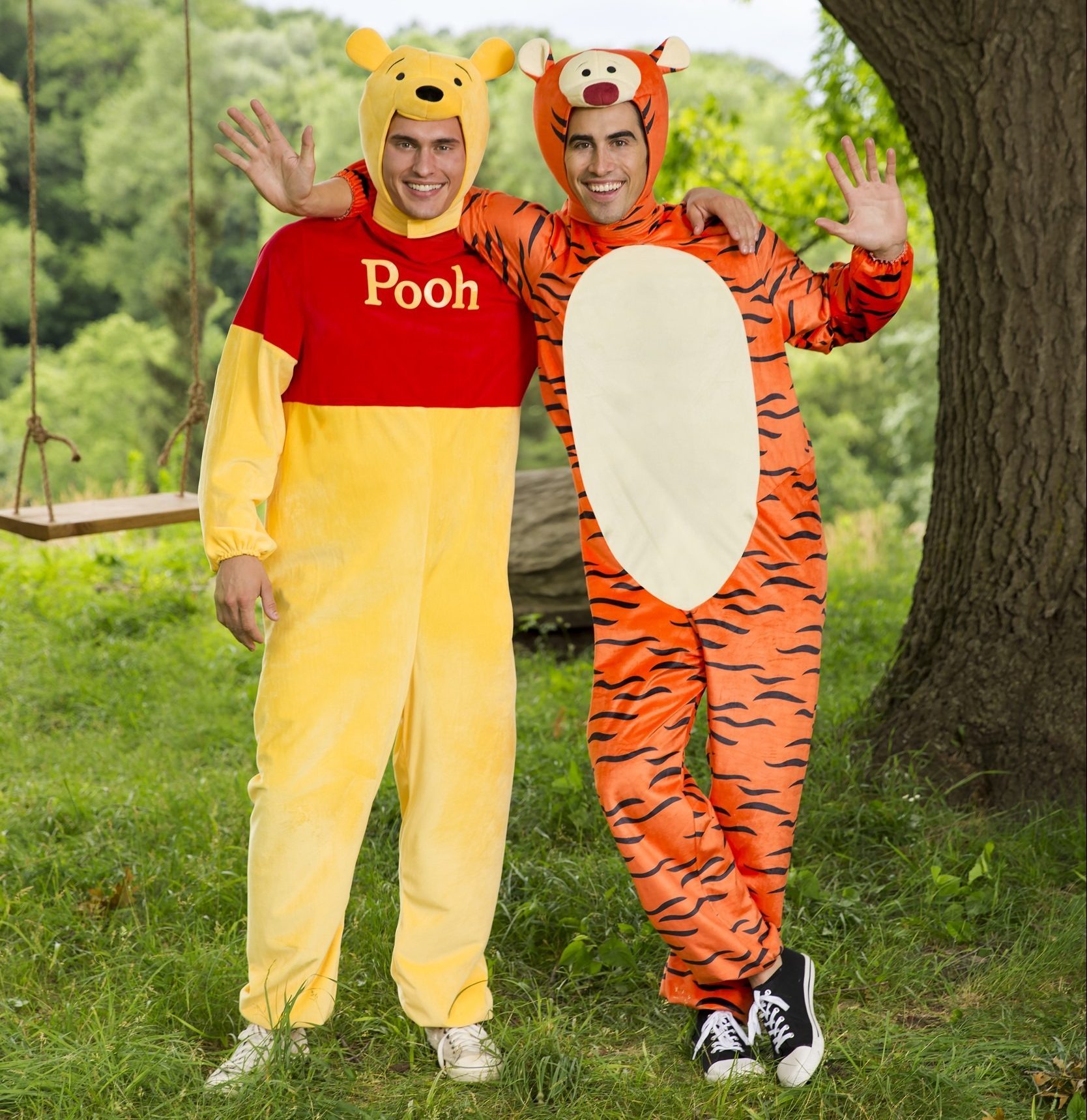 Two people wear Winnie the Pooh and Tigger costumes, waving and smiling, standing in a grassy area with trees and a wooden swing.