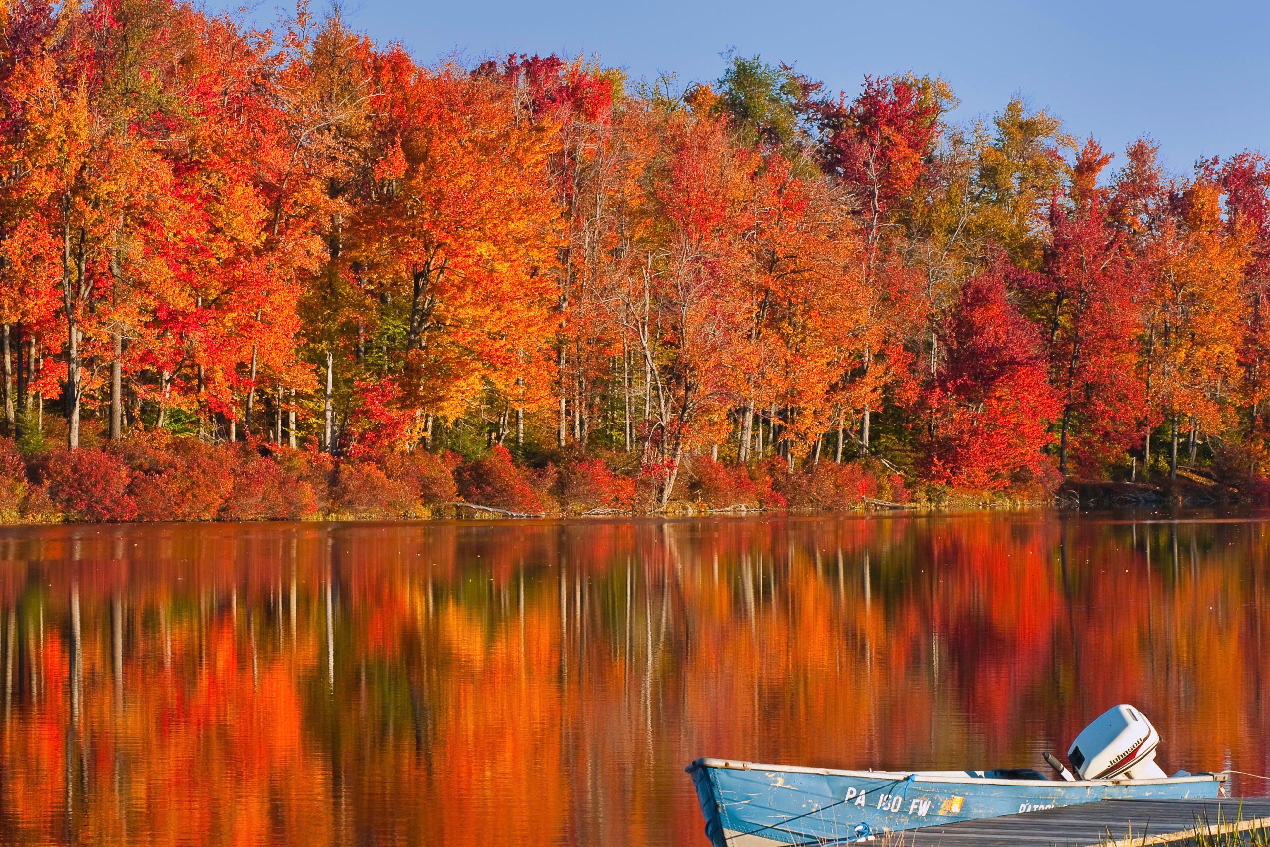 A boat rests on a calm lake, reflecting vibrant autumn trees with red and orange leaves under a clear blue sky.