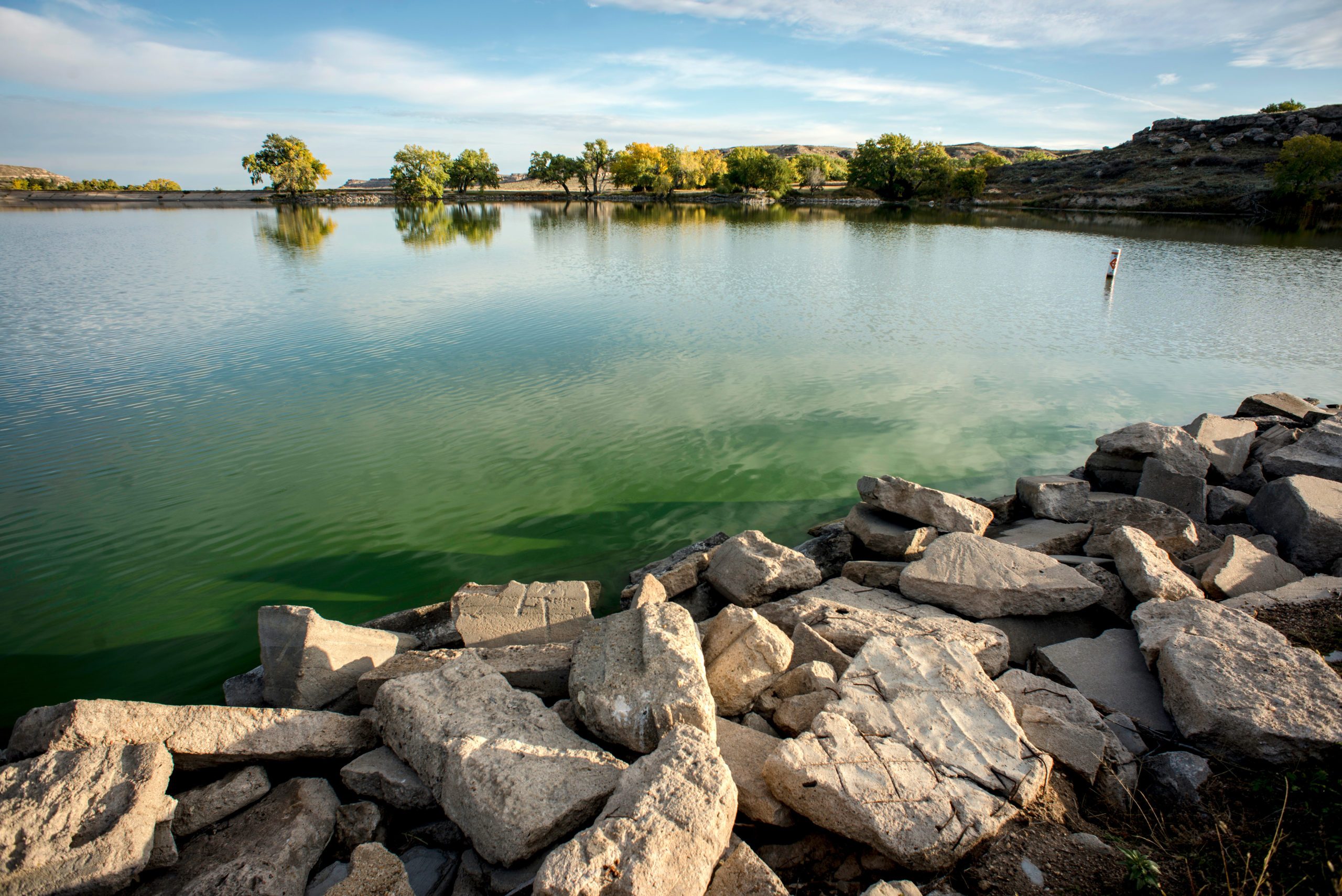 Rocks rest along a tranquil lake with green reflections, surrounded by distant trees and a clear blue sky.