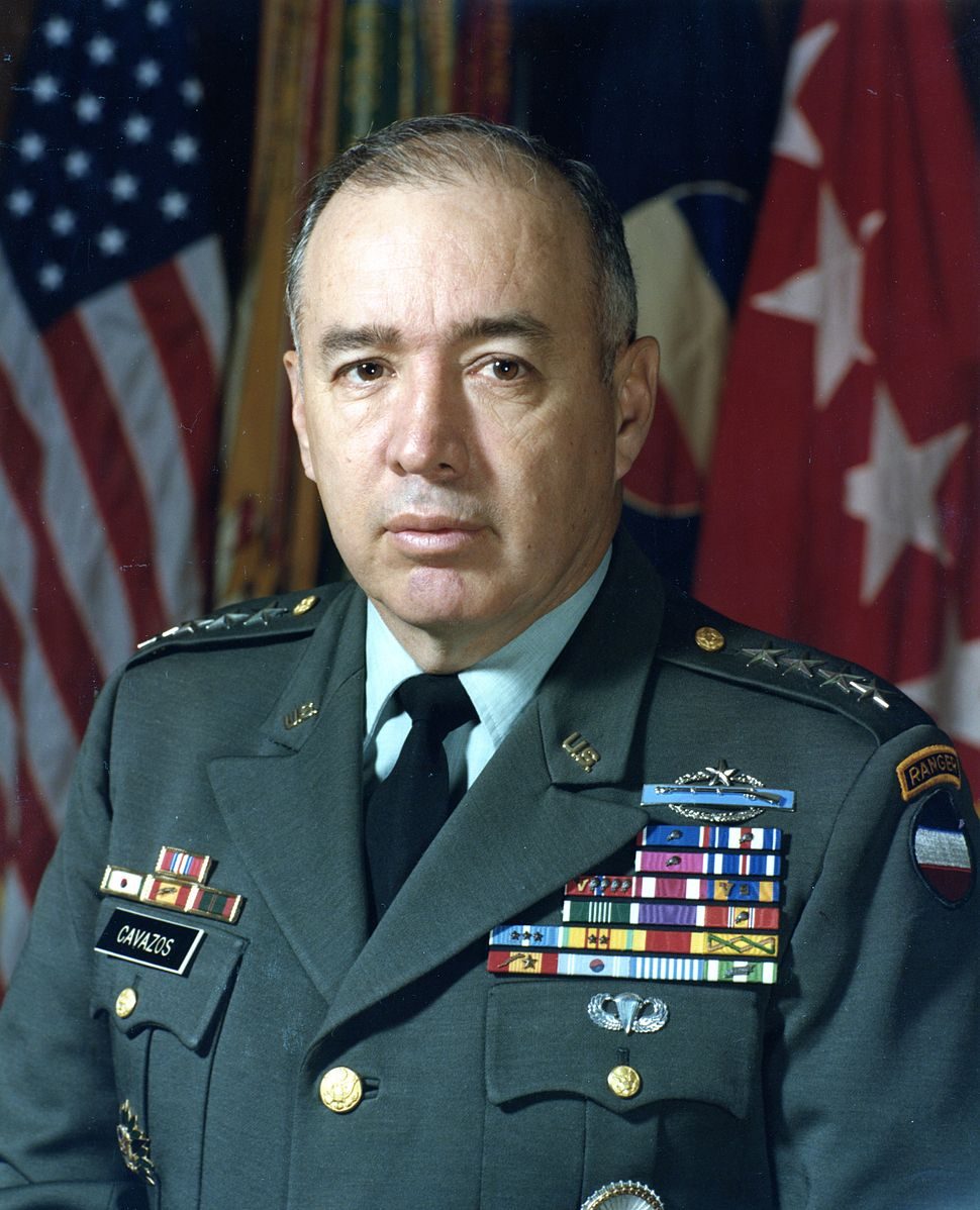 Military officer wears decorated uniform, standing in front of flags.