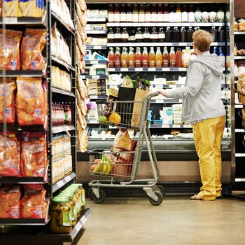 Person pushes shopping cart, selecting items from shelves stocked with various packaged goods, in a grocery store aisle.