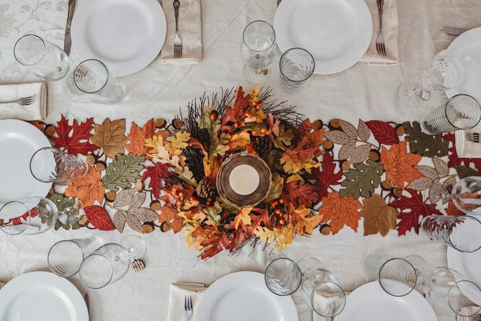 Centerpiece candle sits amidst fall leaves on a dining table, surrounded by plates, glasses, and utensils on a white tablecloth.