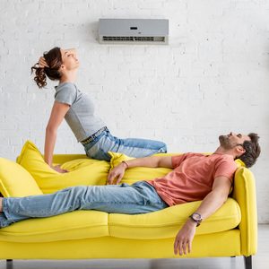 Two people relax on a yellow couch beneath a wall-mounted air conditioner in a white, minimalist living room with shelves holding plants and books.