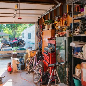 A cluttered garage contains tools and bicycles, opening to a driveway with a white truck and a parked RV under shaded trees.