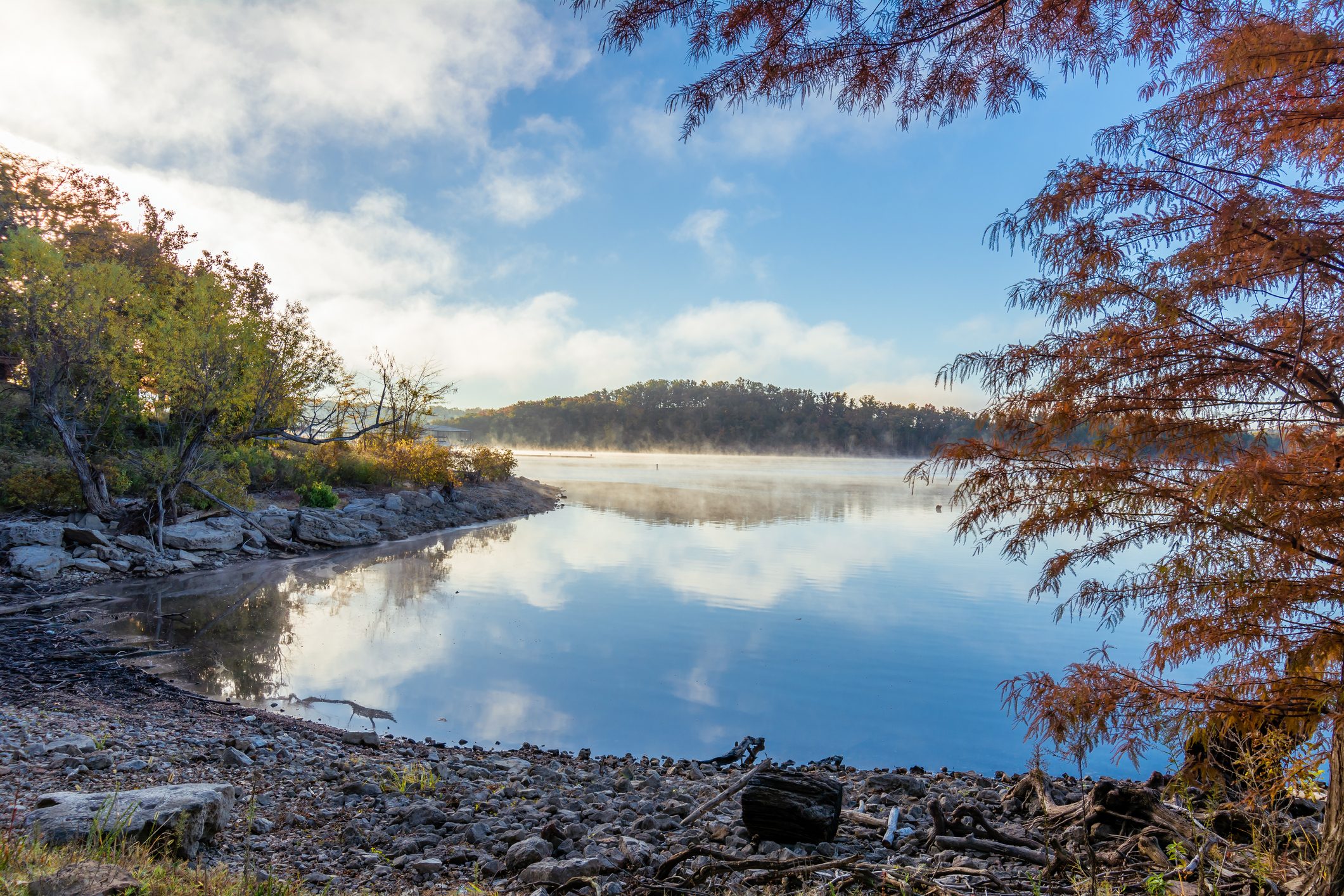 Calm lake reflects clouds, surrounded by autumn trees and rocky shore, under a clear blue sky.