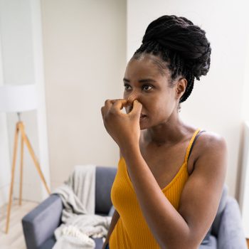 Woman pinching nose, standing in a living room with a gray couch, lamp, and blanket in the background.