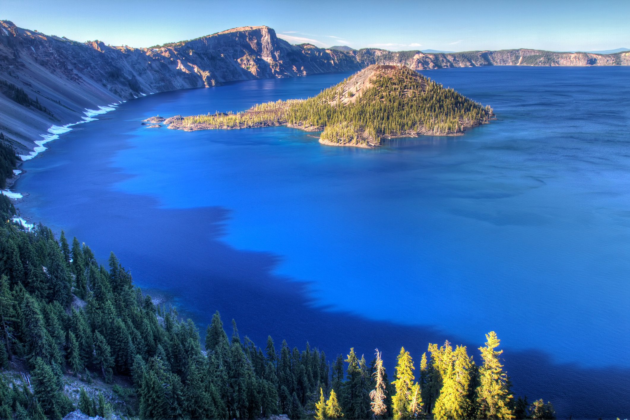 Island covered in trees surrounded by deep blue lake, nestled within steep rocky cliffs, under a clear sky.
