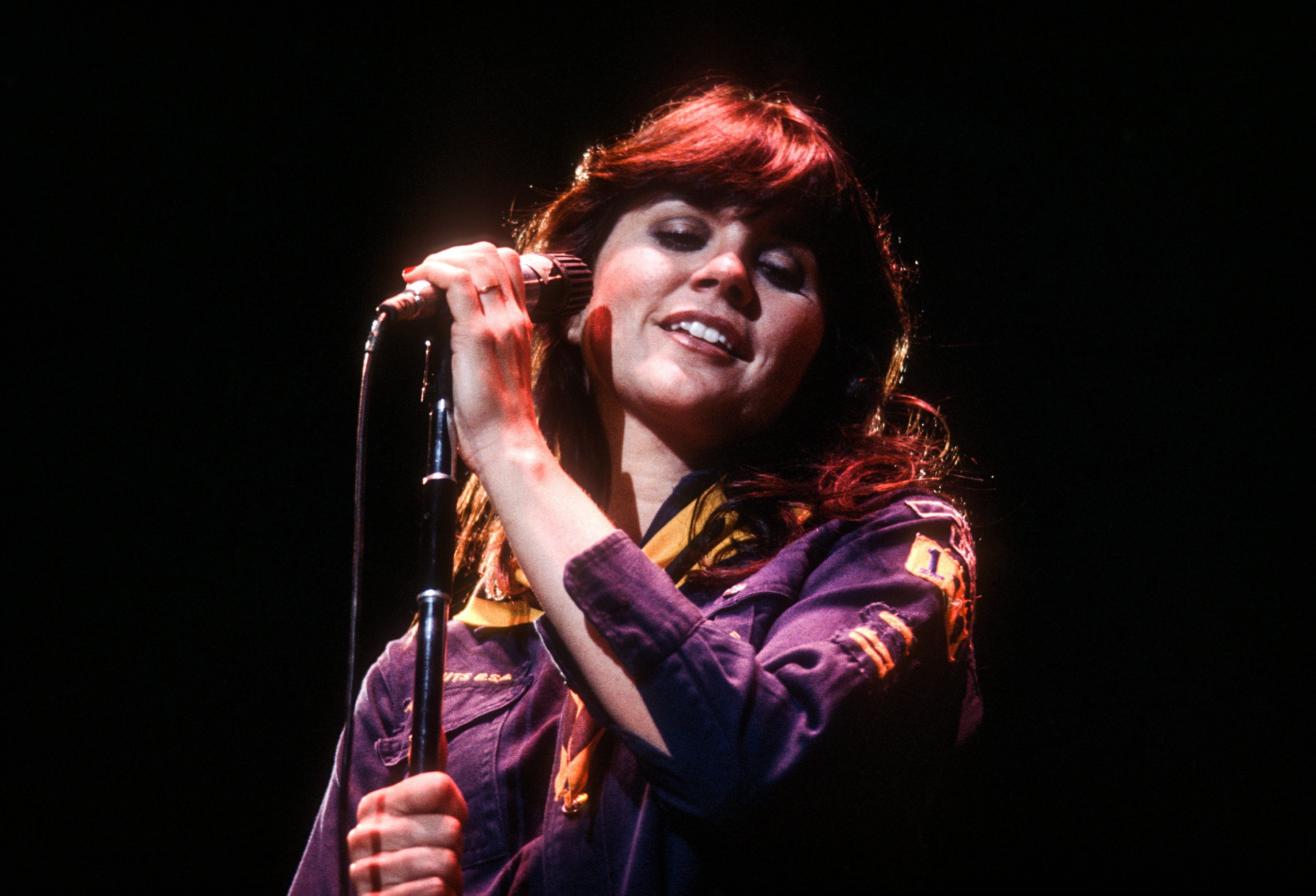 Singer smiles while holding a microphone, performing on a dimly lit stage, wearing a jacket with patches.