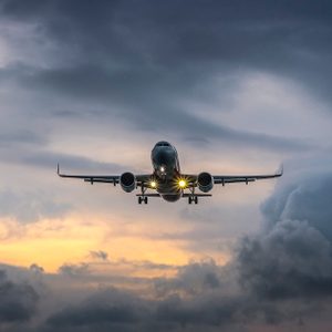 Airplane approaches with landing lights on, set against a cloudy, dramatic sky at dusk.