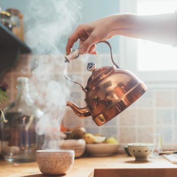 Copper kettle pours steaming water into a cup on a wooden kitchen countertop with jars and plants in the background.