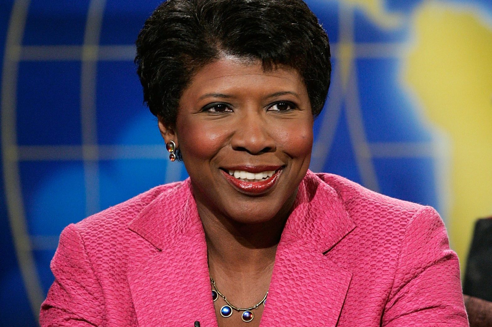 A person smiles, wearing a pink jacket and necklace, against a blue and yellow background with abstract shapes.
