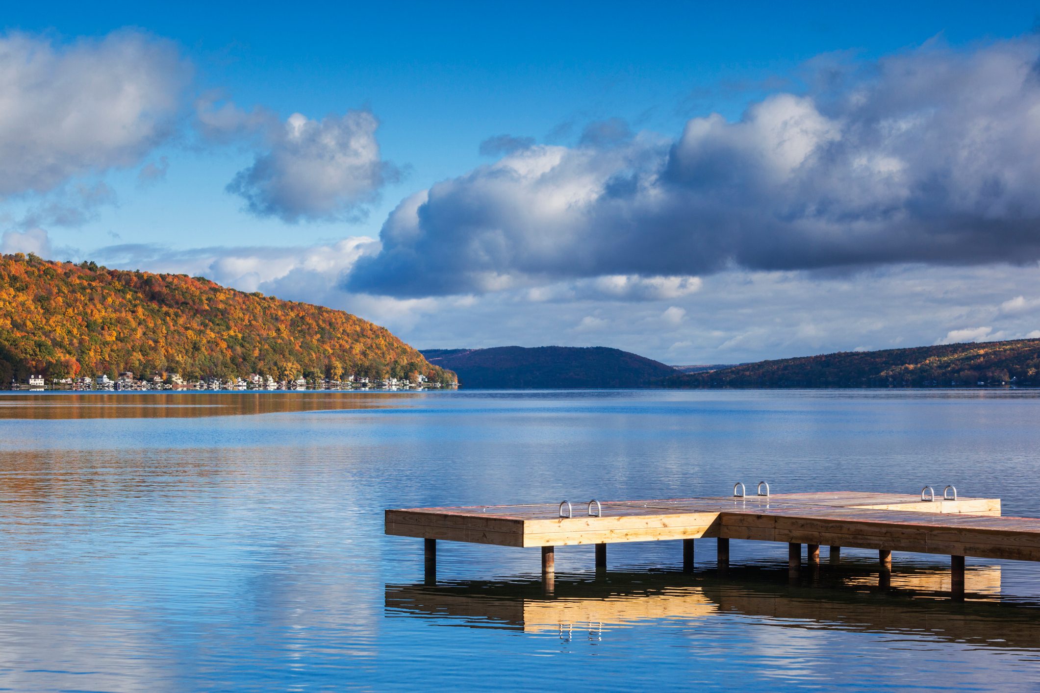 Wooden dock extends over calm lake; autumn-colored hills and scattered houses in the background, under a blue sky with fluffy clouds.