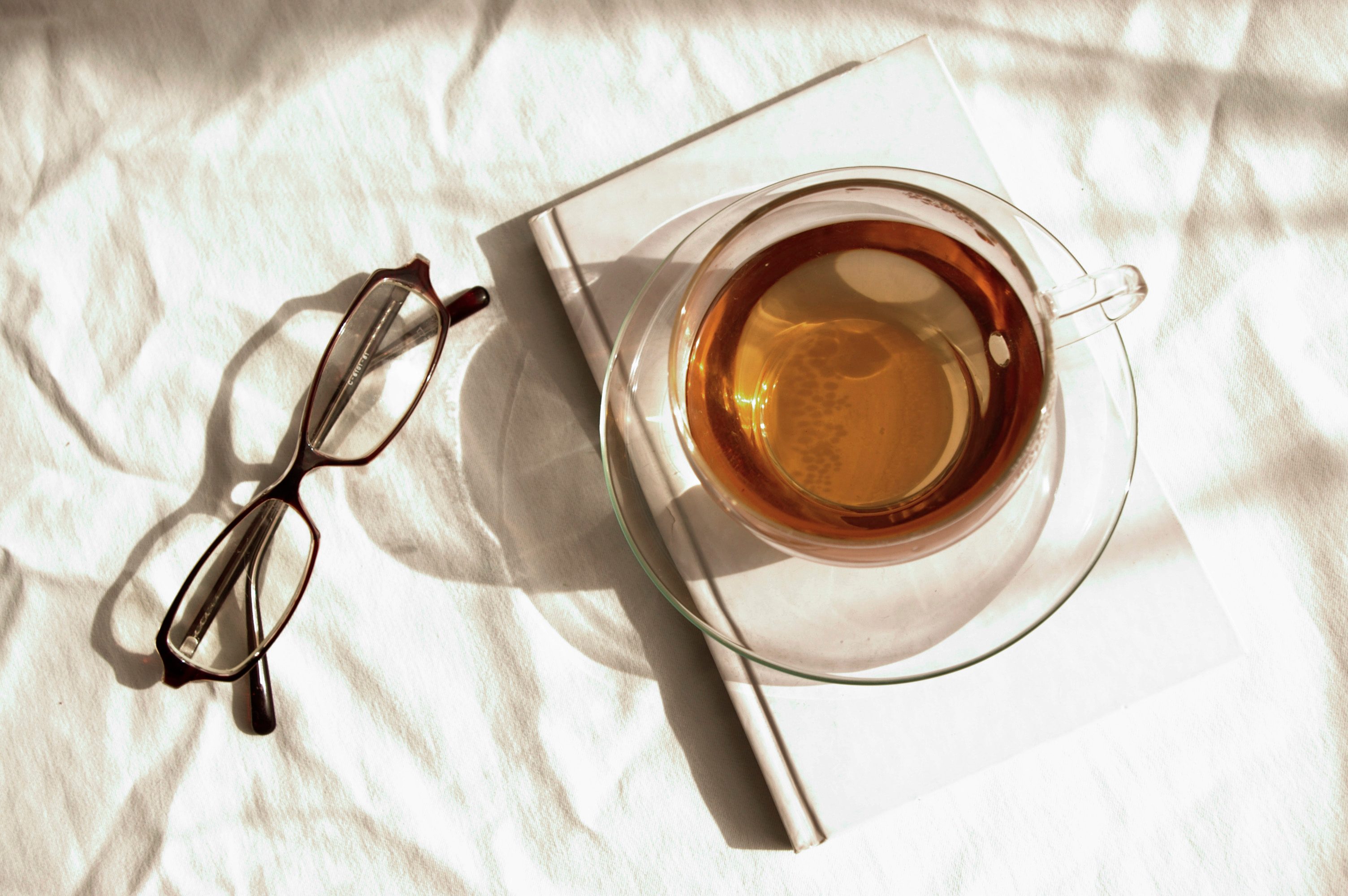 A teacup rests on a closed book, beside eyeglasses, illuminated by soft sunlight on a textured white surface.