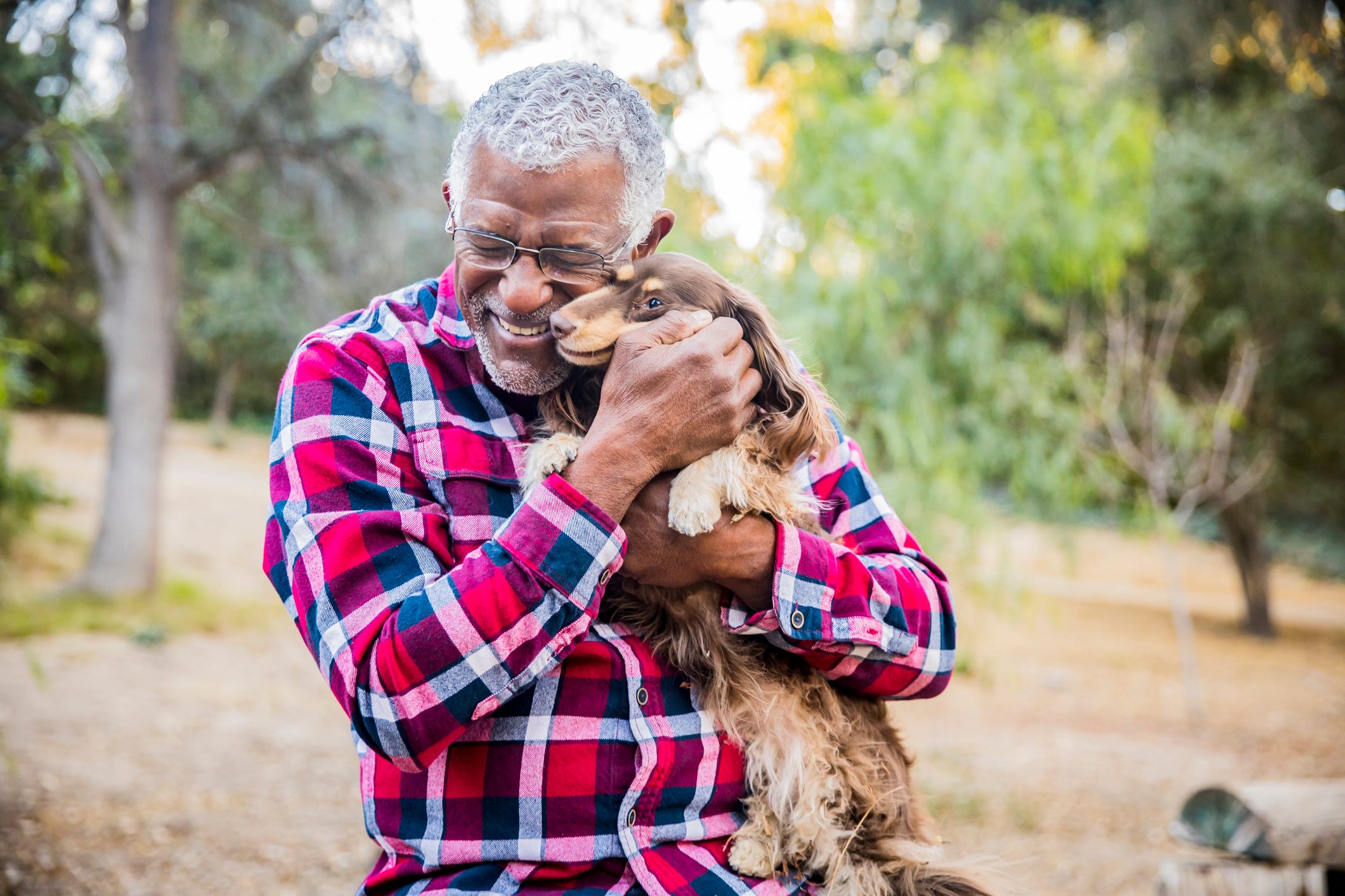 Man embraces small dog affectionately, smiling broadly, in a park with trees and greenery surrounding them.