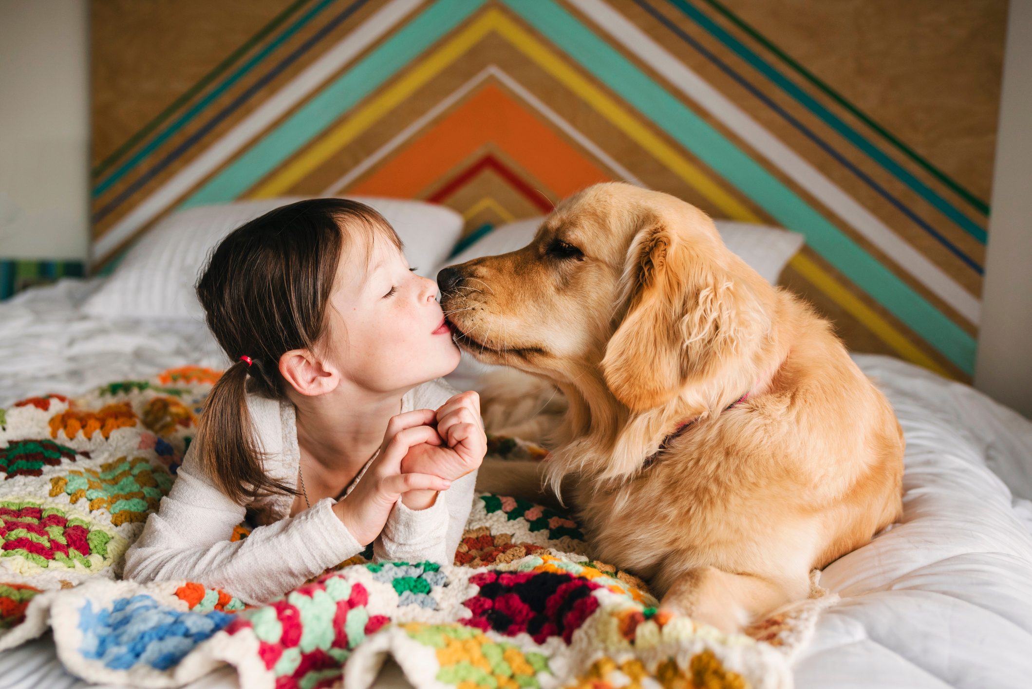 Child kisses golden retriever on a bed, under a colorful, crocheted blanket; geometric wall pattern in background.