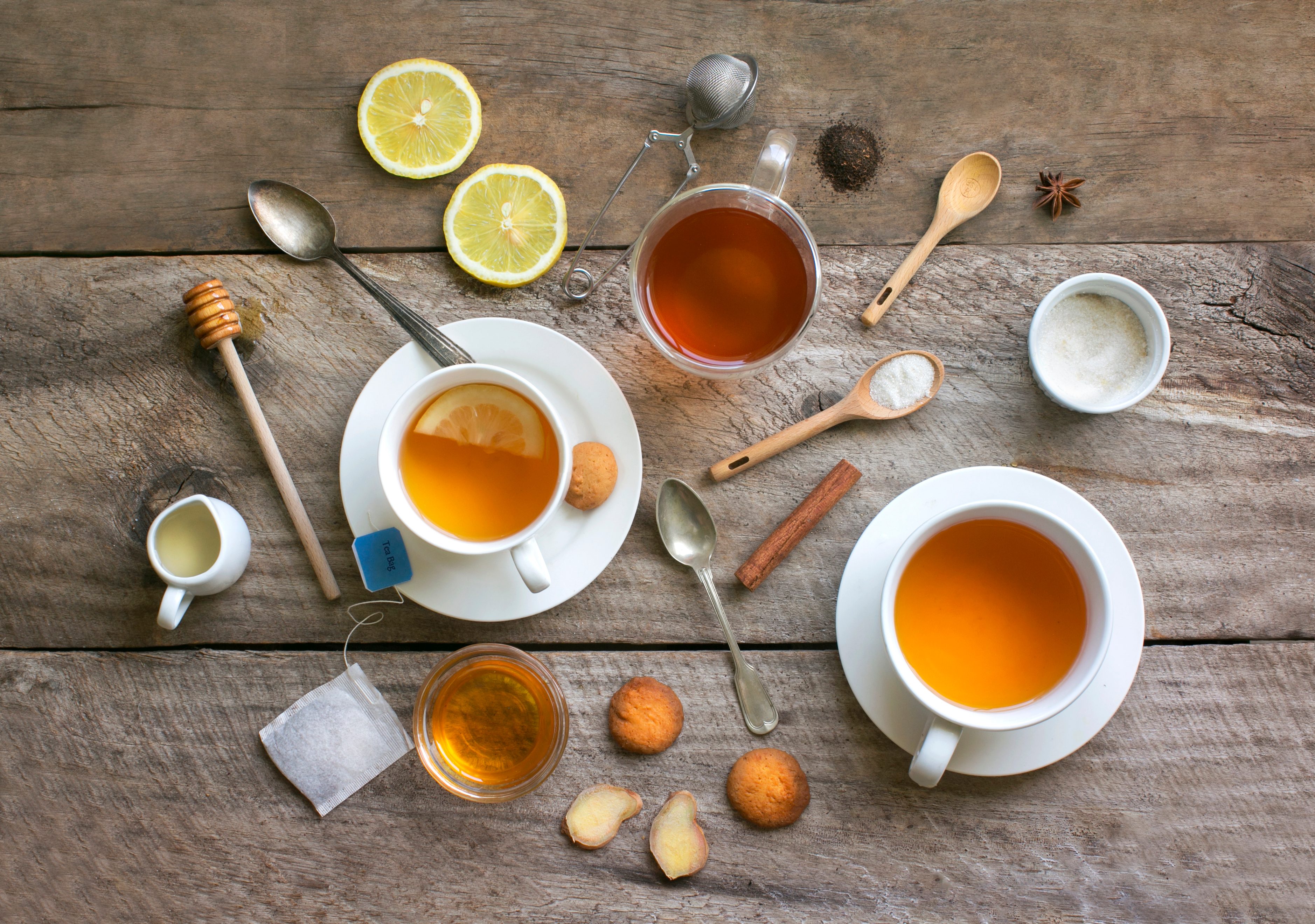 Tea cups sit on a wooden table with lemon slices, honey, cookies, and spoons arranged casually.