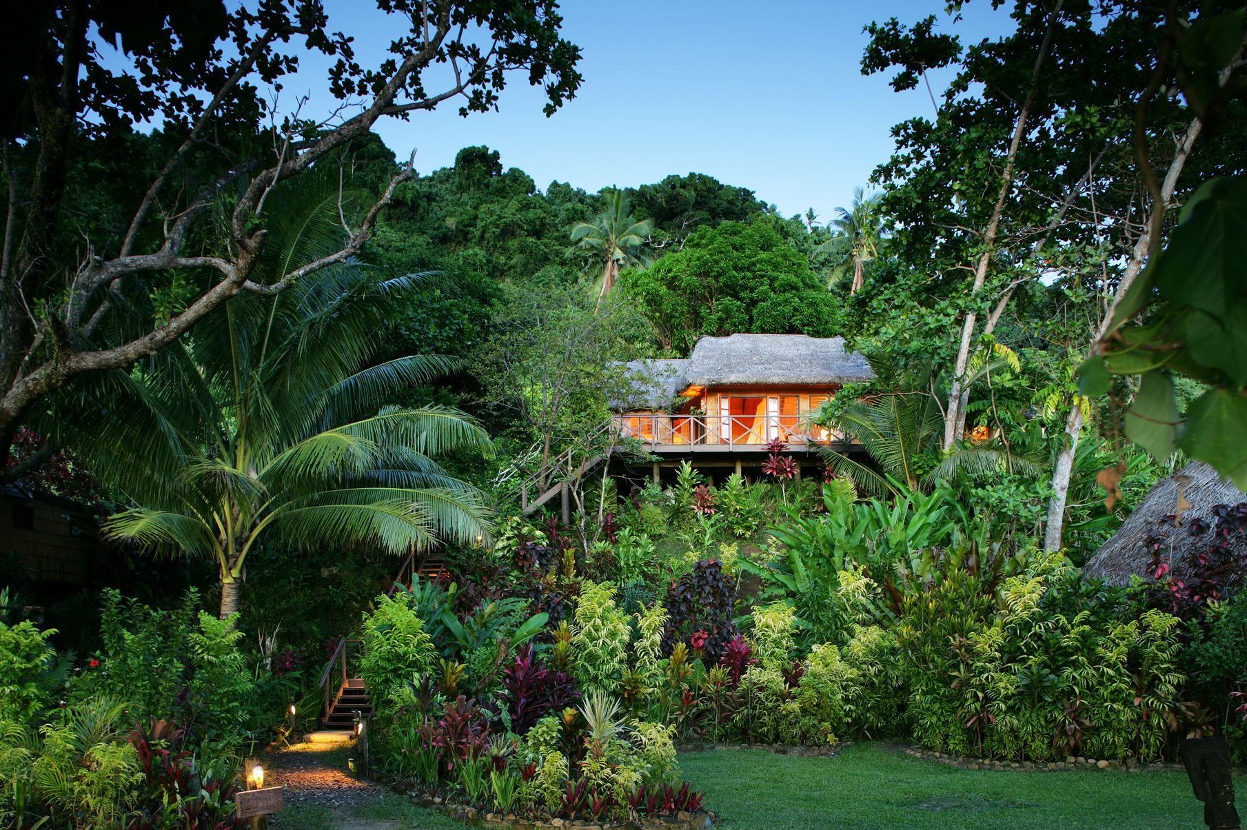 A thatched-roof house sits nestled within lush, tropical vegetation and vibrant plants, with a garden pathway winding through dense greenery leading toward it.