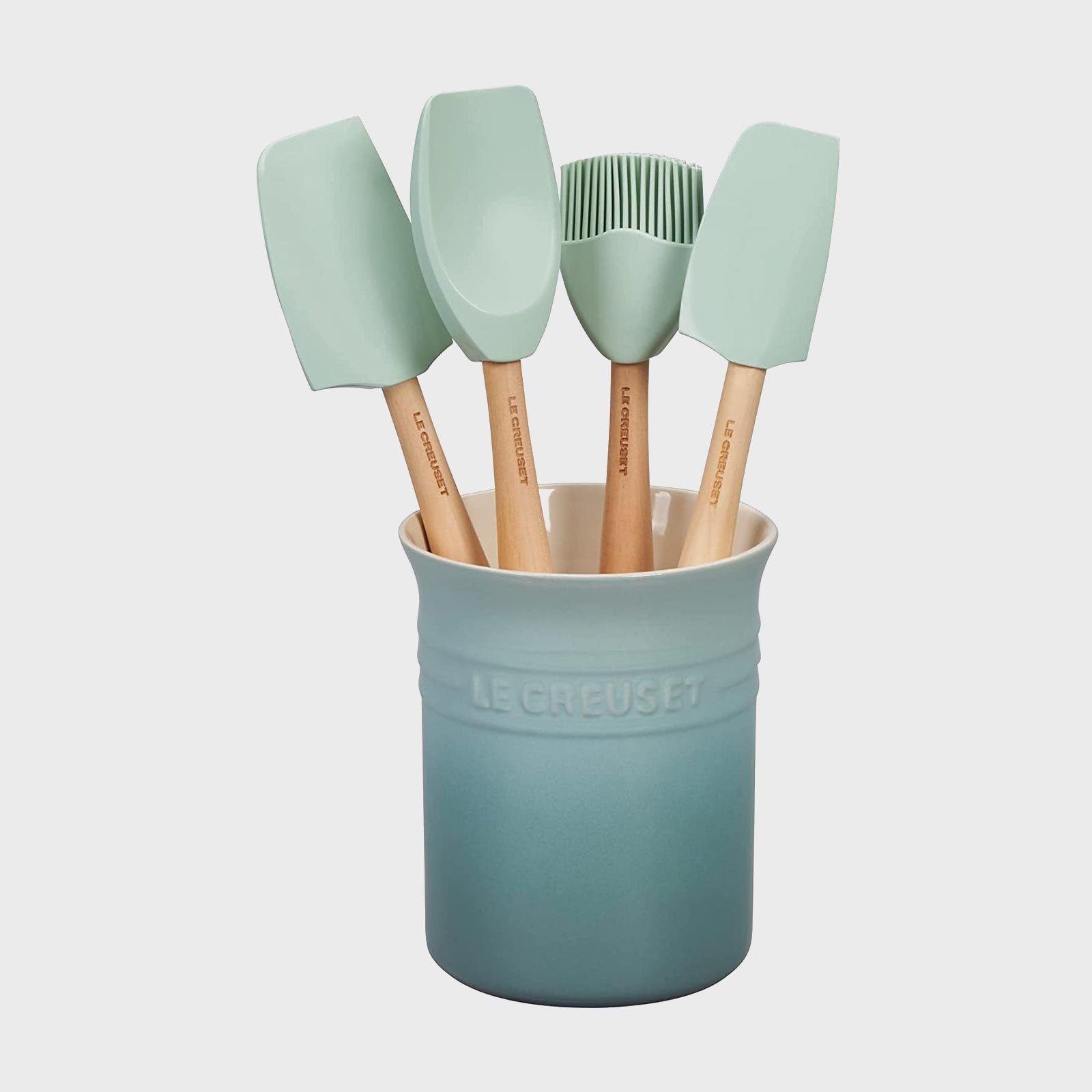 Ceramic container holds four wooden-handled silicone utensils in pastel colors, labeled "Le Creuset," against a plain background.