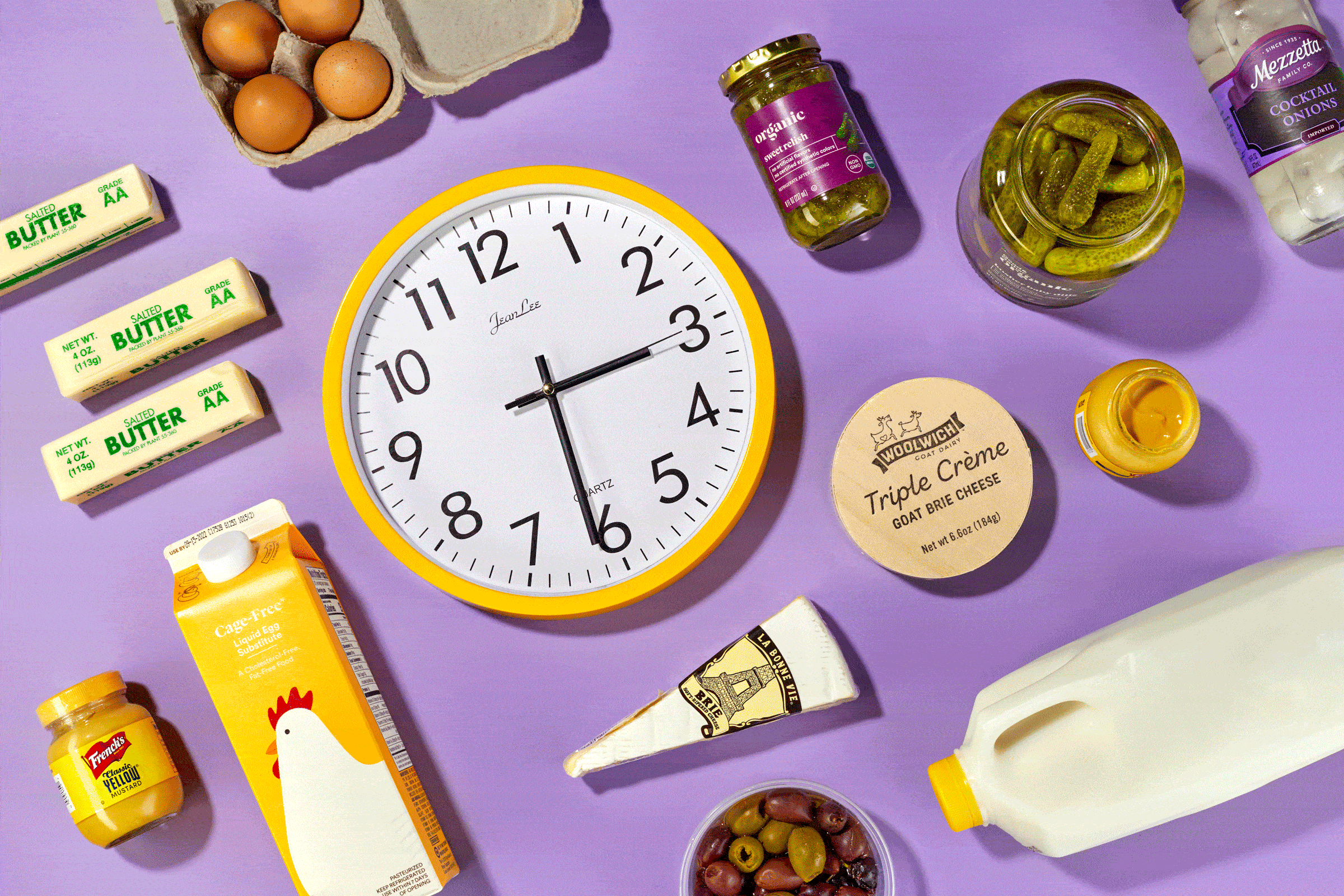 Clock displaying time, surrounded by various foods like eggs, butter, relish, pickles, cheese, mustard, milk, and olives, on a purple background.